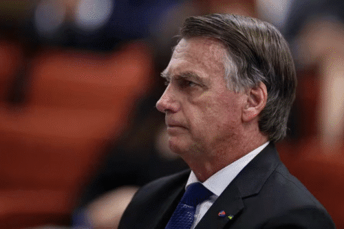Imagem do Bolsonaro, de cabelo grisalho, vestindo terno e gravata azul, em um ambiente formal, com expressão séria, possivelmente durante uma audiência ou reunião.