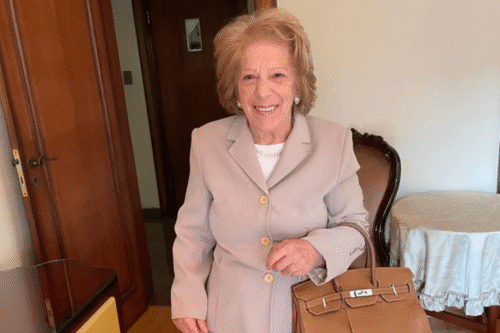 Maria Berklian sorridente com cabelo pintado, vestido bege e bolsa de mão enquanto está em ambiente interno