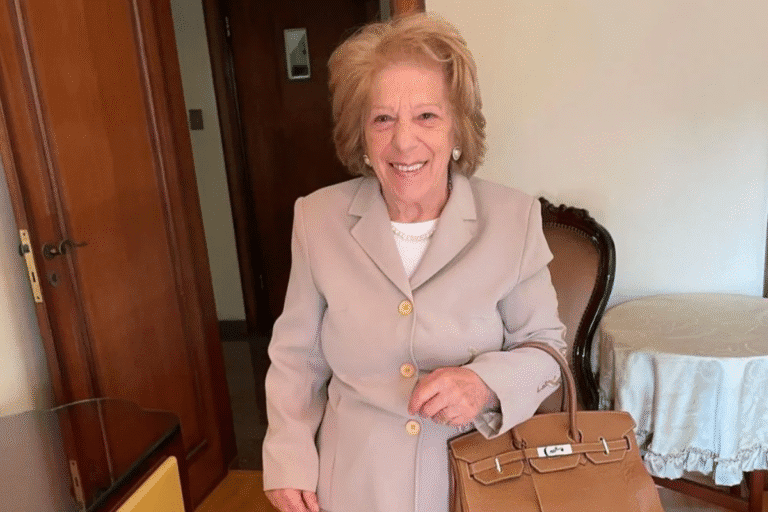 Maria Berklian sorridente com cabelo pintado, vestido bege e bolsa de mão enquanto está em ambiente interno