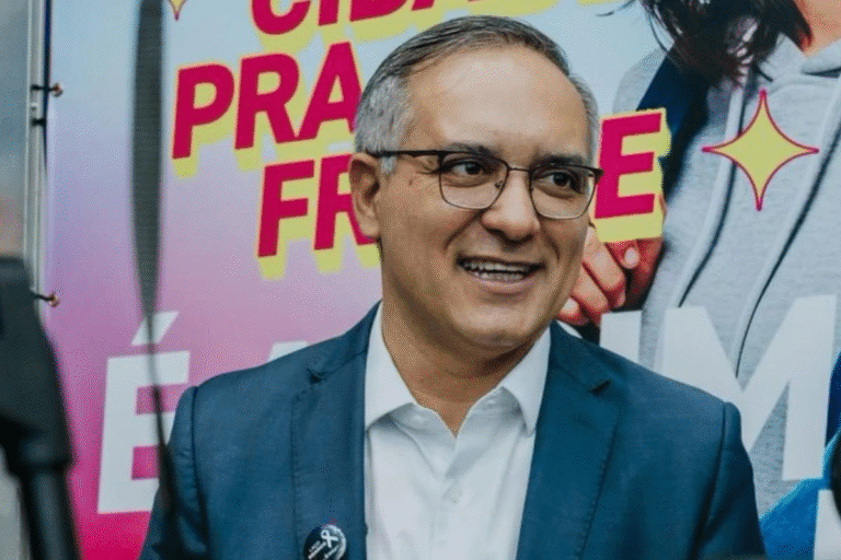 Prefeito de São Bernardo do Campo de terno azul, com sorriso, usando óculos, em ambiente de evento político com fundo colorido e mensagens de campanha