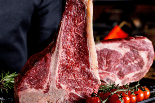 Imagem de um corte de carne bovina, com textura suculenta e marmorizada, preparado para churrasco, decorado com tomates cereja e ramos de alecrim.