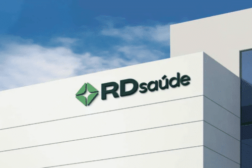 Imagem de um edifício moderno com o logo da RD Saúde, uma clínica ou centro de saúde, sob um céu azul com algumas nuvens, destacando-se na arquitetura branca.