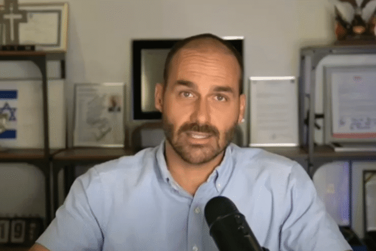 Eduardo Bolsonaro na frente de computadores, com ambiente de escritório ao fundo, usando camisa de botão azul claro.