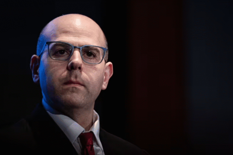 Stephen Miran sério com óculos, usando terno e gravata, em uma fonte de luz dark, sem sorriso, em cenário escuro