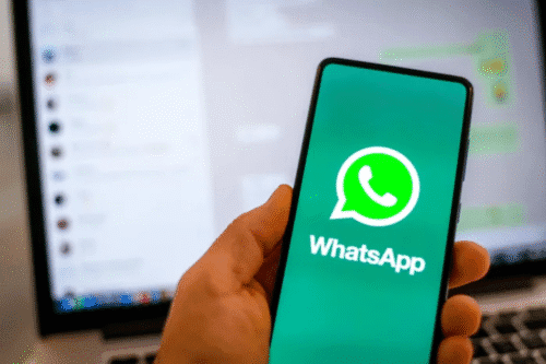 Pessoa segurando um smartphone exibindo o logo do WhatsApp com uma tela de computador ao fundo.