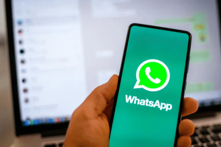Pessoa segurando um smartphone exibindo o logo do WhatsApp com uma tela de computador ao fundo.
