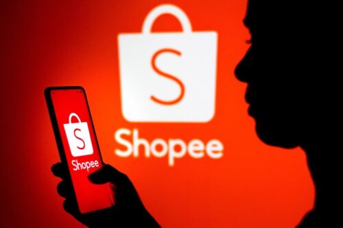 Pessoa acessando o app da Shopee, com o símbolo da marca ao fundo.