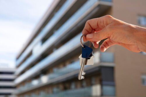 Mão segurando uma chave de apartamento em frente a um prédio de apartamentos, simbolizando aluguel ou compra de imóveis.