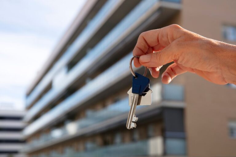 Mão segurando uma chave de apartamento em frente a um prédio de apartamentos, simbolizando aluguel ou compra de imóveis.