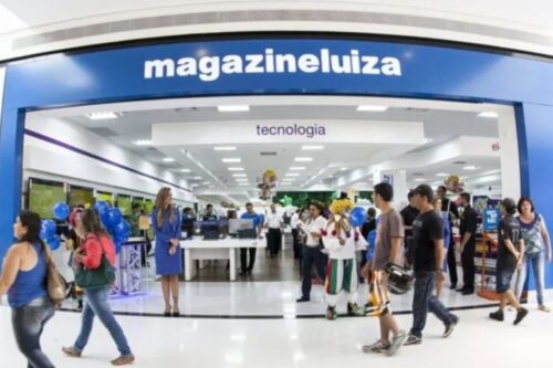 Pessoa entrando na loja MagazineLuiza especializada em tecnologia, com clientes e funcionários dentro e fora do estabelecimento, decorado com balões e sinalização azul.