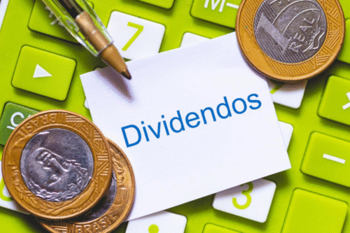 Imagem de moedas e teclado com uma nota escrita 'Dividendos', simbolizando retorno financeiro de investimentos ao lado de uma calculadora.