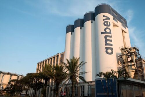Fábrica da Ambev com silos altos e logotipo destacado, cercada por palmeiras e estrutura industrial em um dia ensolarado