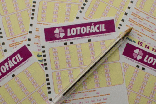 Uma caneta repousa sobre vários bilhetes de loteria "Lotofácil" com grades e números, dispostos em um esquema de cores amarelo e magenta,