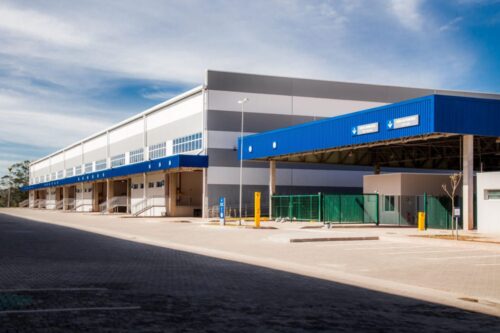 BTLC11: Imagem de um centro de distribuição ou armazém moderno com fachada azul e detalhes em cinza, localizado em um ambiente com céu parcialmente nublado.