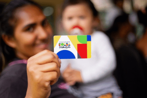 Pessoa segurando cartão do programa 'Famiília Família' em evento, com outras pessoas ao fundo, em um ambiente social e alegre.