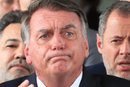 Bolsonaro de terno com expressão facial de dúvida ou preocupação, cercado por outras pessoas ao fundo.