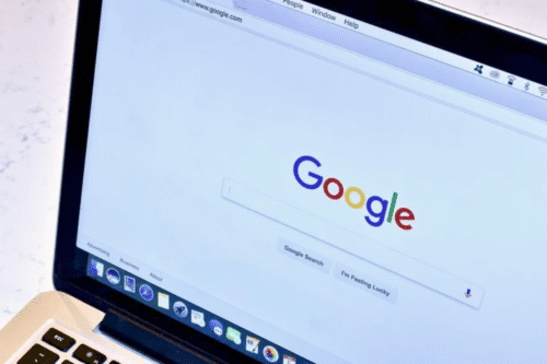 Tela de computador exibindo a página inicial do Google, com o logotipo colorido no centro da tela. Ao lado, uma parte do teclado do computador portátil.