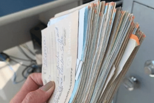 Pessoa segurando um grande monte de cheques, com aspecto de documentos financeiros, em um ambiente interno.