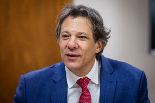 Haddad cin roupa social azul, com expressão séria, em ambiente interno de escritório, representando liderança e profissionalismo.