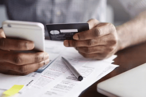 Pessoa realizando uma operação de pagamento com cartão de crédito e celular, em uma mesa com documentos e papel calculating.