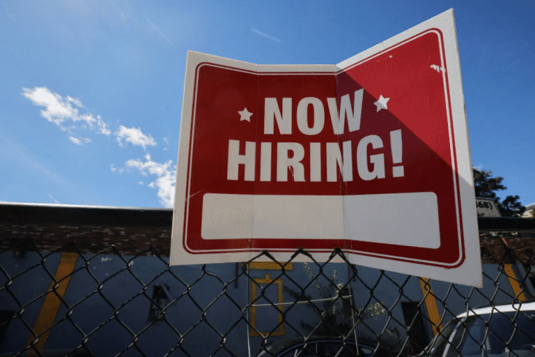 Cartel de 'Now Hiring!' pendurado em uma cerca de arame, anunciando vaga de emprego disponível no momento.