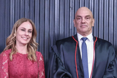 Viviane de Moraes com um vestido de renda vermelha sorri ao lado de Alexandre de Moraes com um robe preto formal com detalhes vermelhos. O fundo apresenta painéis escuros verticais, criando um ambiente formal.