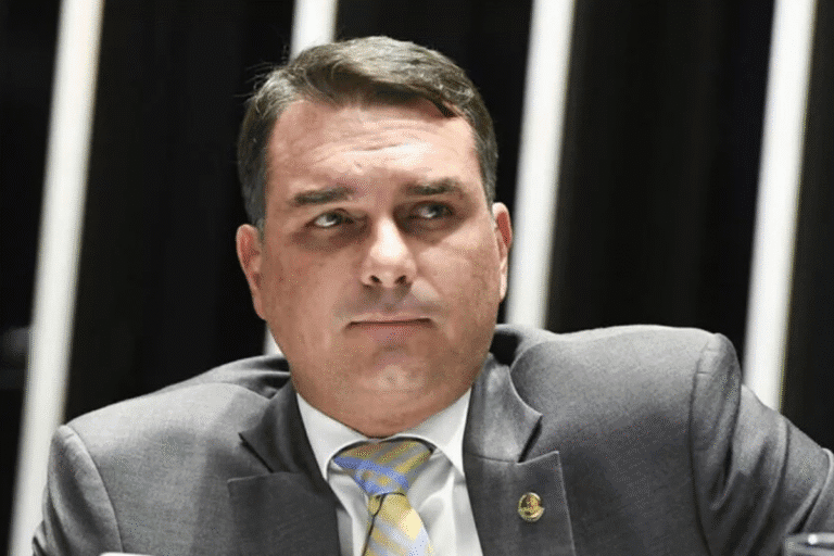 Flávio Bolsonaro de terno cinza com gravata listrada amarela está sentado em frente a faixas verticais de luz. Ele parece pensativo e sério.