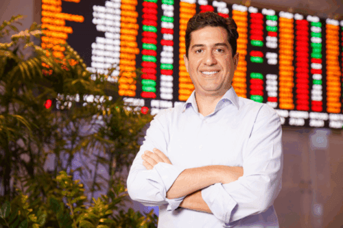 Foto de Felipe Paiva, direto da B3, em frente a um gráfico de ações.