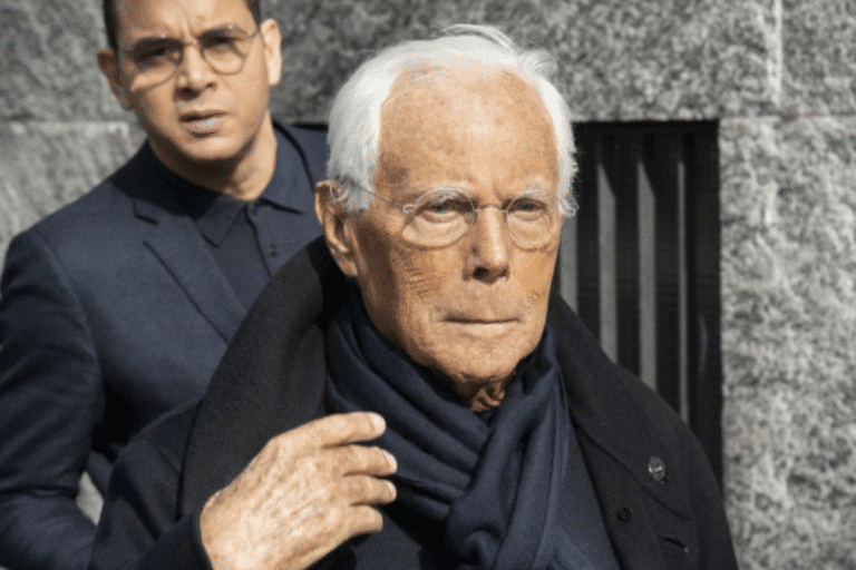 Imagem de Giorgio Armani, usando óculos e um sobretudo preto, acompanhado por um homem mais jovem ao fundo, ambos em um ambiente urbano de pedra.