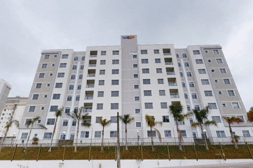 Imagem de um edifício residencial moderno com várias janelas, localizado em uma área com árvores palmadas, sob céu nublado.