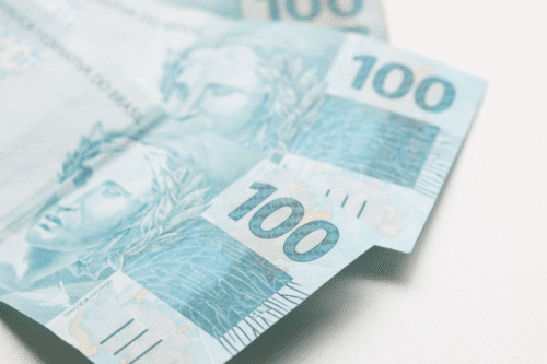 Notas de 100 reais dispostas em uma superfície branca, destacando a linha e detalhes do dinheiro brasileiro.