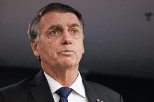 Bolsonaro sério e pensativo, usando terno escuro e gravata, com cabelo grisalho, possivelmente um político ou executivo, em fundo neutro.