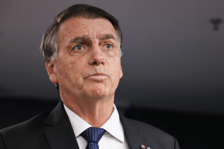 Bolsonaro sério e pensativo, usando terno escuro e gravata, com cabelo grisalho, possivelmente um político ou executivo, em fundo neutro.
