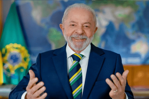 Imagem de um homem idoso sorridente, usando terno azul e gravata listrada, em um ambiente com bandeira do Brasil ao fundo, simbolizando liderança e governança.