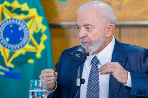 Imagem de Lula, usando terno azul, falando em um microfone durante uma entrevista ou reunião, com a bandeira do Brasil ao fundo.