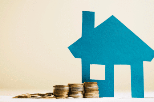 Imagem de uma casa em papel azul ao lado de pilhas de moedas, simbolizando investimento em imóveis e planejamento financeiro.