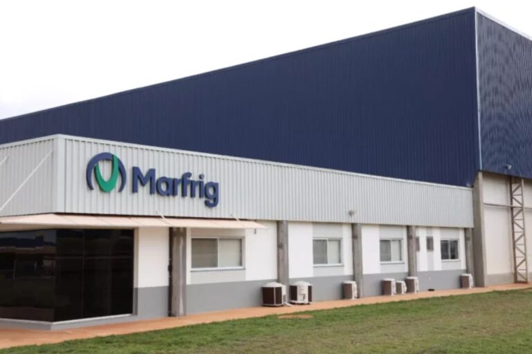 Fachada da fábrica da Marfig, empresa de fabricação de alimentos, com logotipo visível, parede de metal azul e branca, e área verde na frente.