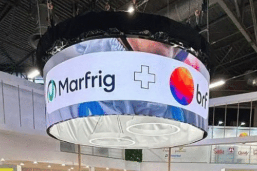Display digital uniforme com logotipos de Marfrig e Orfi em um ambiente de feira ou evento de negócios. A tela está suspensa no teto com iluminação do ambiente ao fundo.