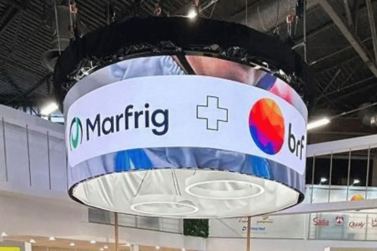 Display digital uniforme com logotipos de Marfrig e Orfi em um ambiente de feira ou evento de negócios. A tela está suspensa no teto com iluminação do ambiente ao fundo.