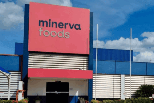 Imagem da fachada do Minerva Foods, com um letreiro em destaque na cor rosa e azul, sob céu com nuvens brancas e céu azul, mostrando o ambiente externo do estabelecimento.