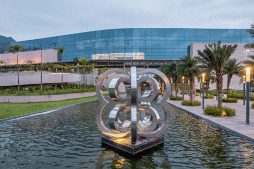 Escultura moderna de metal no centro de um parque com lago, ao lado de um prédio de vidro refletindo o céu, cercado por palmeiras e iluminação urbana.
