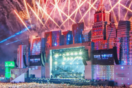 Show ao vivo de um espetáculo de fogos de artifício iluminando o céu sobre uma arena ao ar livre, com palco iluminado e uma cidade ao fundo, proporcionando uma experiência vibrante e animada.