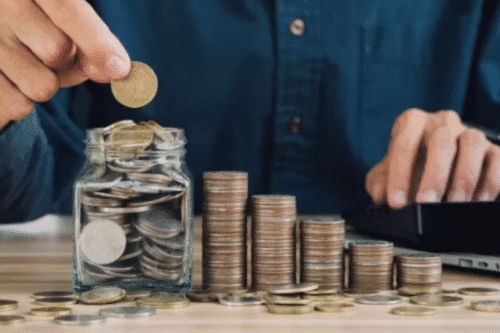 Pessoa colocando uma moeda de ouro em um vidro contendo várias moedas de diferentes tamanhos, ao lado de pilhas de moedas organizadas de maneira crescente, simbolizando economia e investimento.