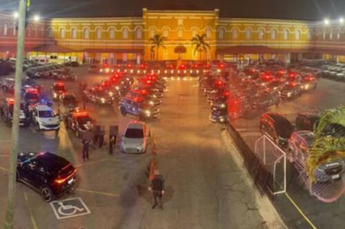 Imagem do estacionamento de uma grande quantidade de veículos, incluindo carros de polícia, com uma estrutura histórica ao fundo, iluminada à noite