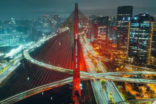 Imagem noturna do Viaduto Santa Paula iluminado na cidade de São Paulo, com o trânsito movimentado e arranha-céus ao fundo, destacando a engenharia e a beleza urbana