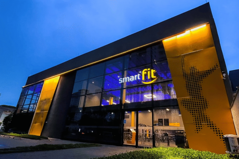 Edifício de academia de ginástica smart fit com fachada moderna de vidro e iluminação amarela à noite, destacando-se na cidade.