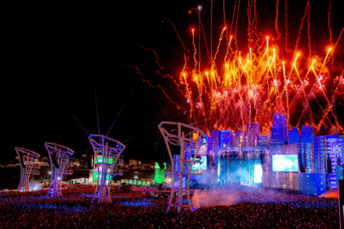 Show de fogos de artifício iluminando o céu durante um festival de música ao ar livre, com cenário futurista e uma multidão animada aproveitando o evento.