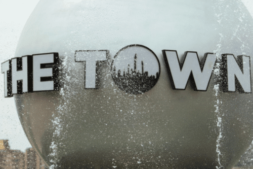 Logotipo de 'The Town' no lado de um prédio, com gotas de água e reflexo no vidro, representando um ponto turístico ou local conhecido, sob um céu nublado.