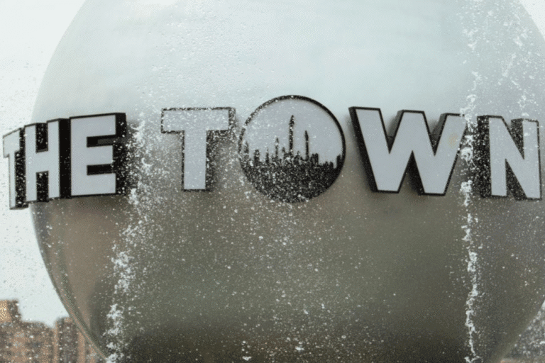 Logotipo de 'The Town' no lado de um prédio, com gotas de água e reflexo no vidro, representando um ponto turístico ou local conhecido, sob um céu nublado.