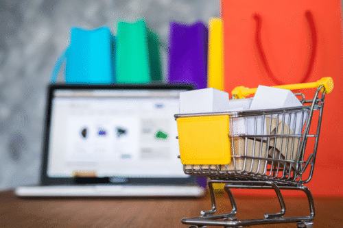 Carrinho de compras em miniatura com caixas de papelão em frente a um laptop e sacolas coloridas ao fundo, simbolizando compras online e vendas pela internet.